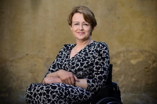 Alt text - Image shows Baroness Tanni Grey-Thompson in a black and white patterned dress sitting in a wheelchair with her arms folded resting on her lap. Tanni has short brown hair swept across her forehead. She is smiling against a yellow sand stone wall background.