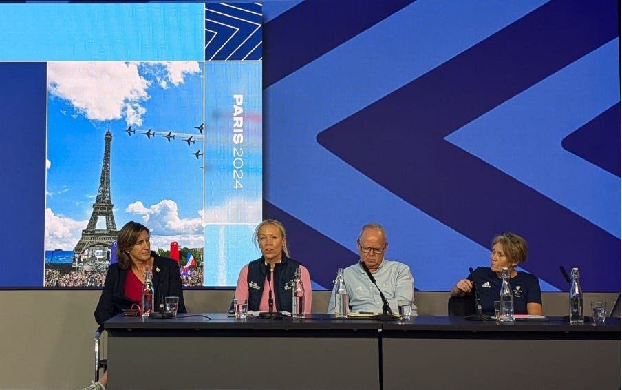 Image shows a panel of 4 people seated behind a desk in conversation. There are individual microphones and bottles placed on the desk. The wall behind has a series of blue arrows as a backdrop and a screen showing a promotional image of Paris 2024 featuring the Eiffel Tower with planes trailing red white and blue smoke.