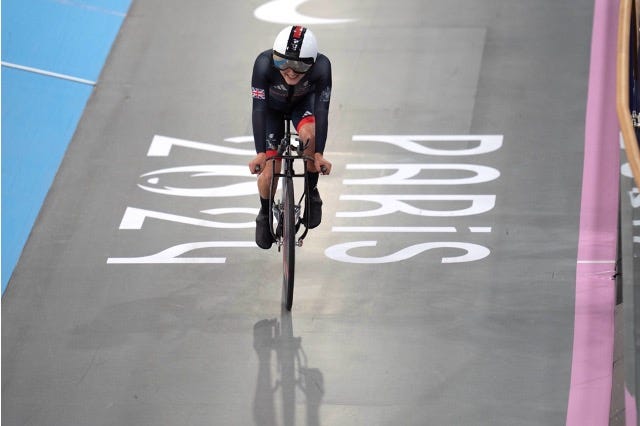 British Paralympian cyclist is racing on the track at the Paris 2024 velodrome, wearing a dark cycling suit and helmet, with "Paris 2024" visible on the track beneath them