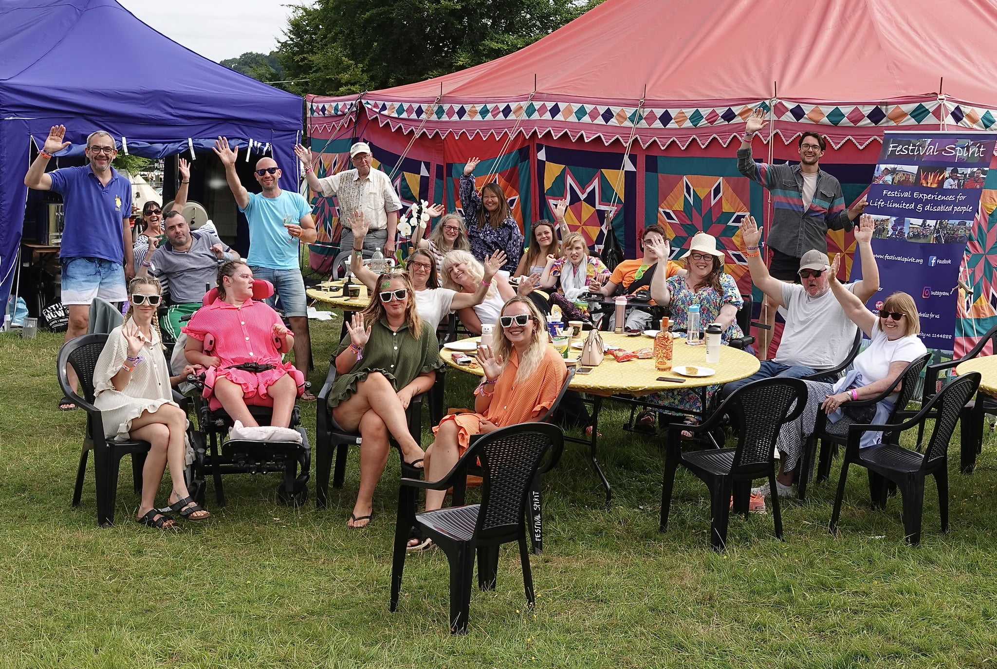 Group of people stood and sat around several tables waving outside purple and red tents infant of a Festival Spirit Banner.