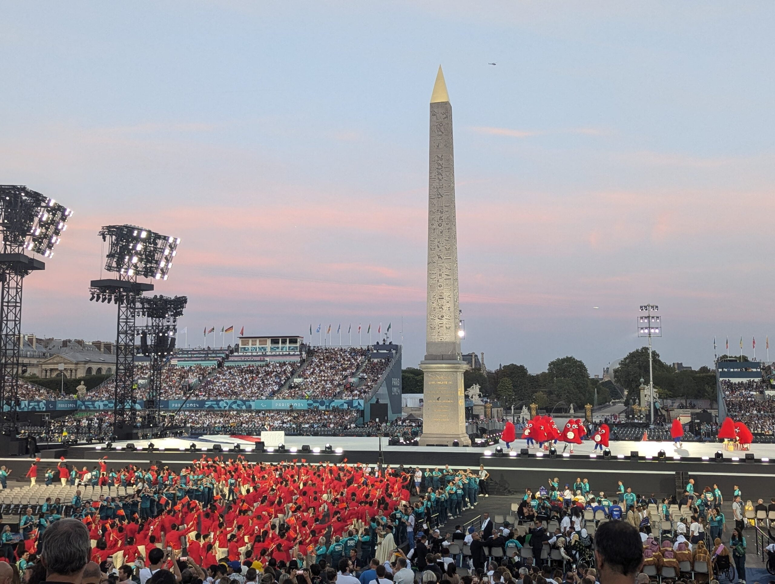 A vibrant scene from the Paris 2024 Opening Ceremony at dusk, featuring a tall obelisk with a golden tip in the center. Surrounding the monument are grandstands filled with spectators, while a large group of performers in red costumes are gathered on the stage. Large lighting rigs illuminate the area, and flags are visible in the background under a pink and blue sky.