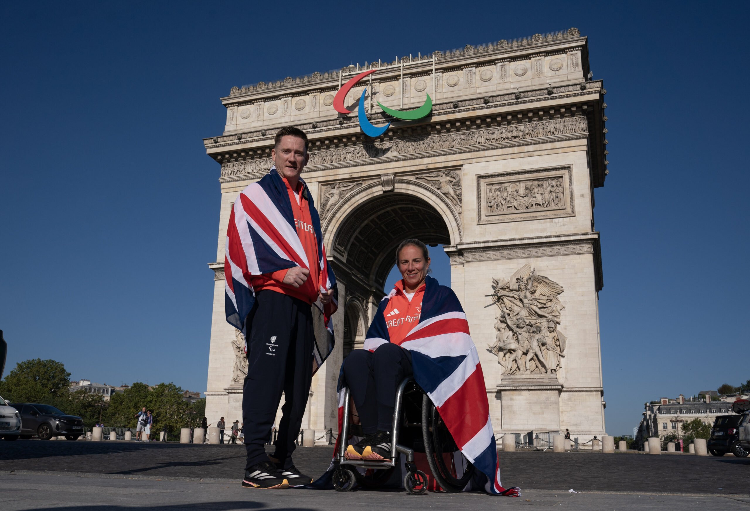 Seven-time Paralympic Wheelchair basketball player Terry Bywater stands next to Wheelchair tennis athlete Lucy Shuker, who will be competing at her fifth Games, seated in a wheelchair. They are both smiling, draped in the Union Jack flag in front of the Arc de Triomphe in Paris wearing Great Britain tracksuits. The Paralympic logo is prominently displayed on the Arc de Triomphe above them, with a clear blue sky in the background.
