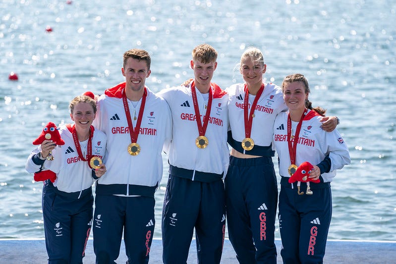 GB Para Rowing Team stand against a water backdrop with their Gold medals. Cox Erin Kennedy, Edward Fuller, Joshua O’Brien, Giedre Rakauskaite and Frankie Allen.  All wearying their GB Team Kit.