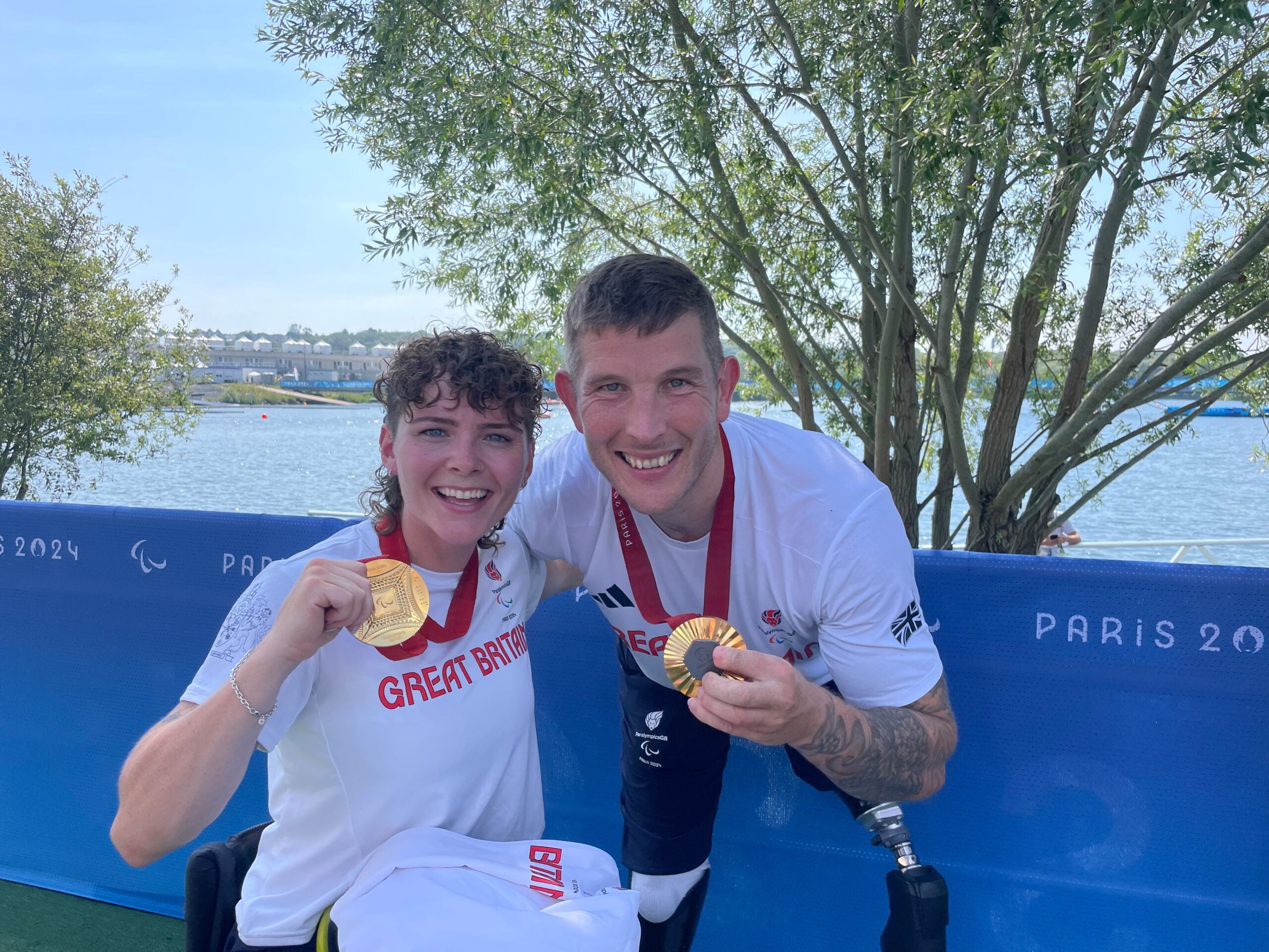 Paralympian Mixed Rowers Lauren Rowles & Gregg Stevenson hold up Gold Medals beaming at the camera. Lauren is seated in a wheelchair with Greg is beside her.  Both have their arms around eachorher
