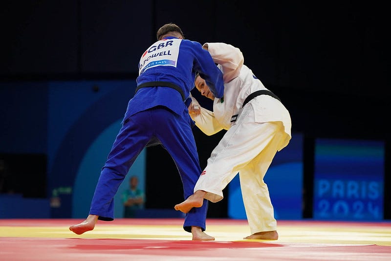 Gold-medal match in the men’s J1 -90kg category at the Paris 2024 Paralympic Games. Daniel Powell from Great Britain, in the blue judogi, is seen battling against Arthur Cavalcante da Silva of Brazil, wearing white. The two athletes are locked in a gripping judo exchange, with the backdrop showcasing the vibrant colors and branding of the Paris 2024 Paralympics.