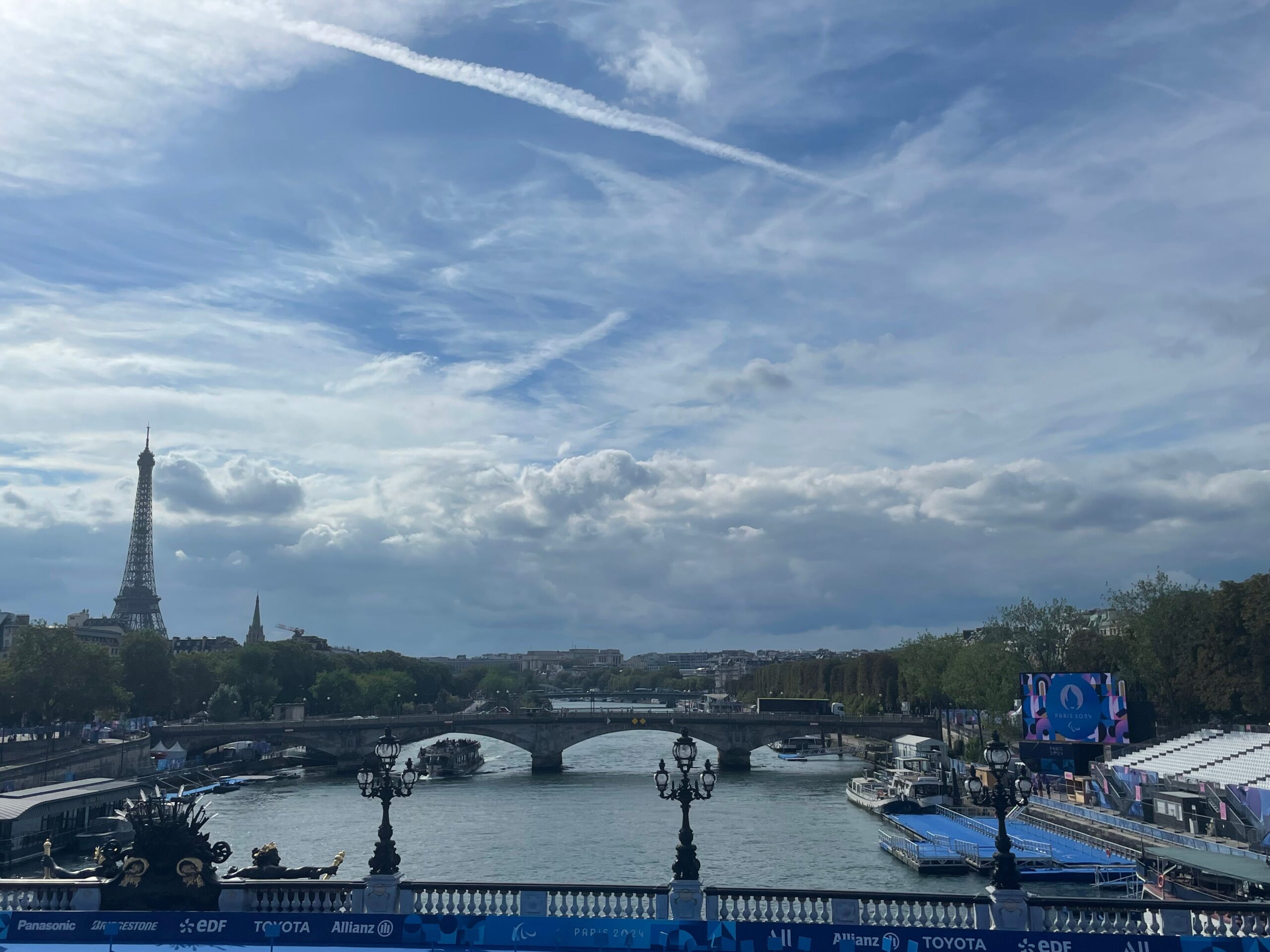 Landscape view from a bridge over the Seine of the Paratriathlon course at the Paris 2024 Paralympic Games. The Eiffel Tower is prominently featured on the left, while a bridge spans the river. On the right, a large screen displays the Paris 2024 Paralympic Games logo, with the triathlon platform lining the riverbank.