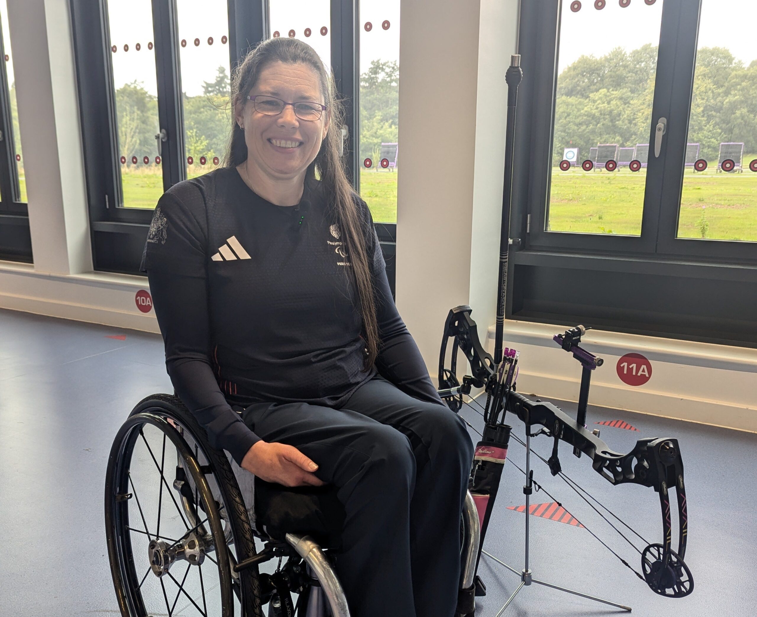 Archer Victoria Kingstone in a wheelchair, wearing a a dark blue ParalympicsGB kit, is smiling with her bow by her side. Behind her our large windows with the a view of a grass archery training area.