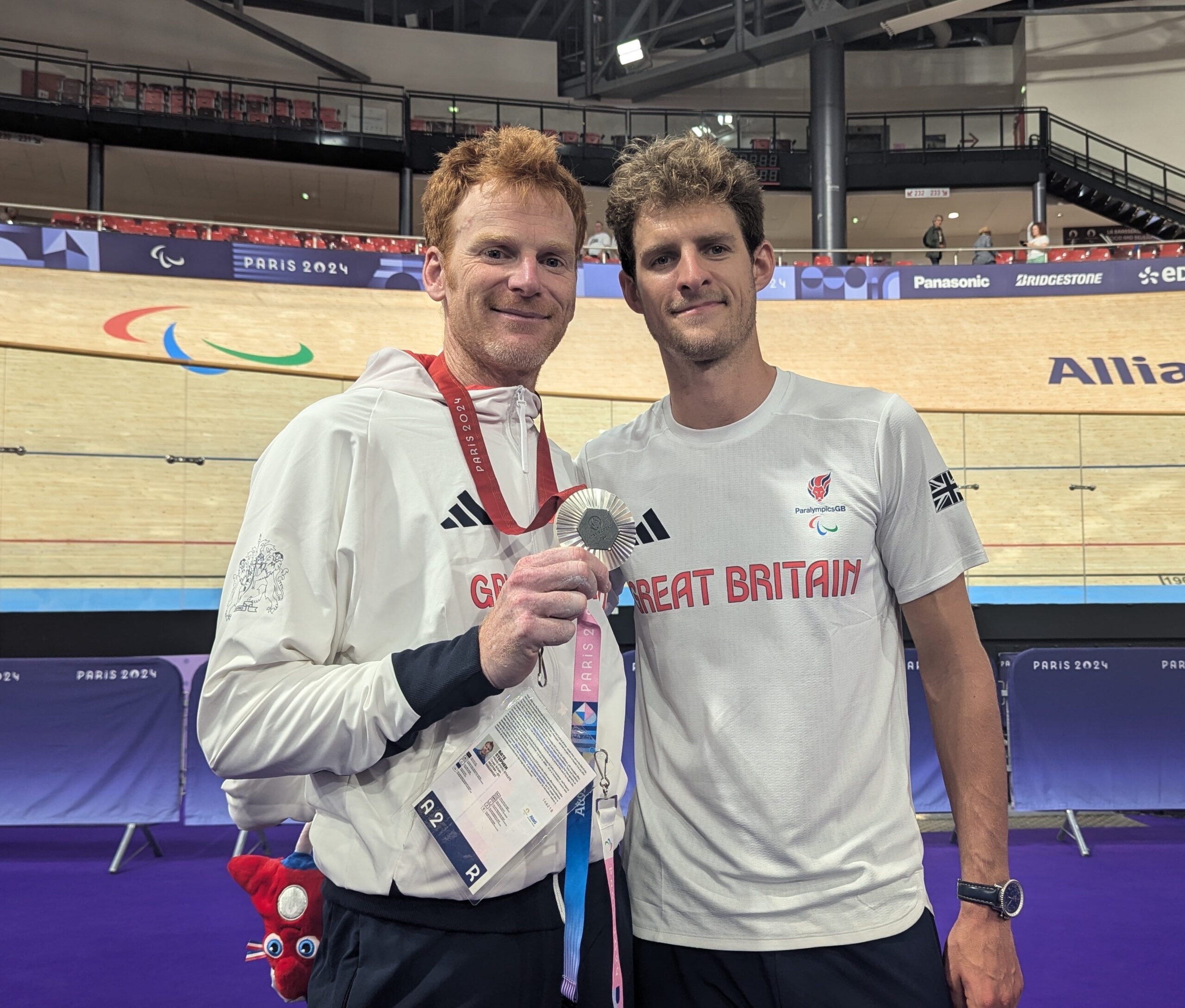 Stephen Bate and Chris Latham, stand together in the velodrome during the Paris 2024 Paralympics. Stephen Bate, wearing a white Great Britain jacket, proudly holds a silver medal. Beside him, Chris Latham, in a white Great Britain T-shirt, smiles. The wooden cycling track and Paralympic banners are visible in the background,.