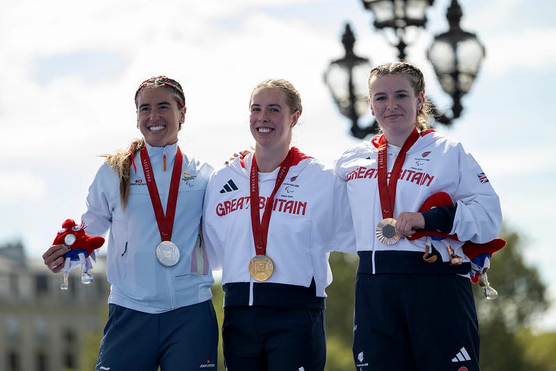 Paralympic triathletes Megan Richter (center) and Hannah Moore (right), along with Spain's Marta Frances Gomes, are pictured smiling with their medals. The background features blurred buildings, trees, an ornate lamppost, and an overcast sky.