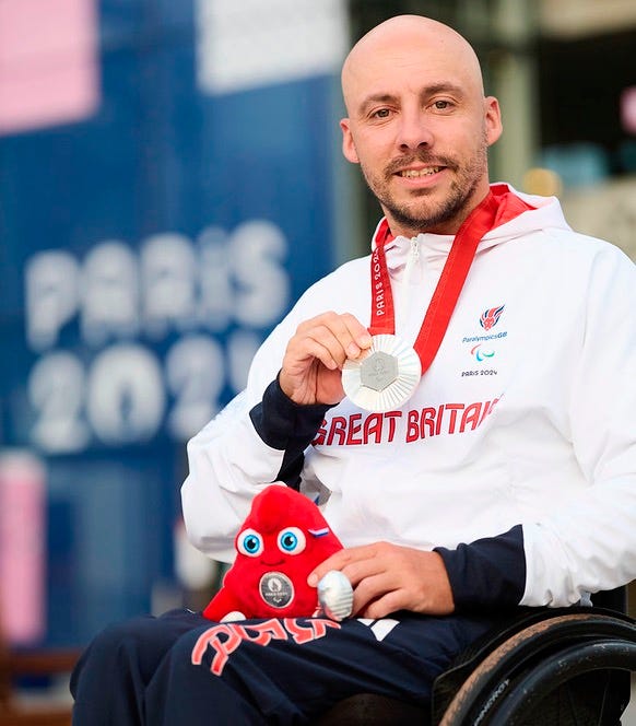 Paralympian Andy Lapthorne in GB kit seated in his wheelchair holding up his Silver Medal and Paralympics Mascot in front of a Paris 2024 banner.
