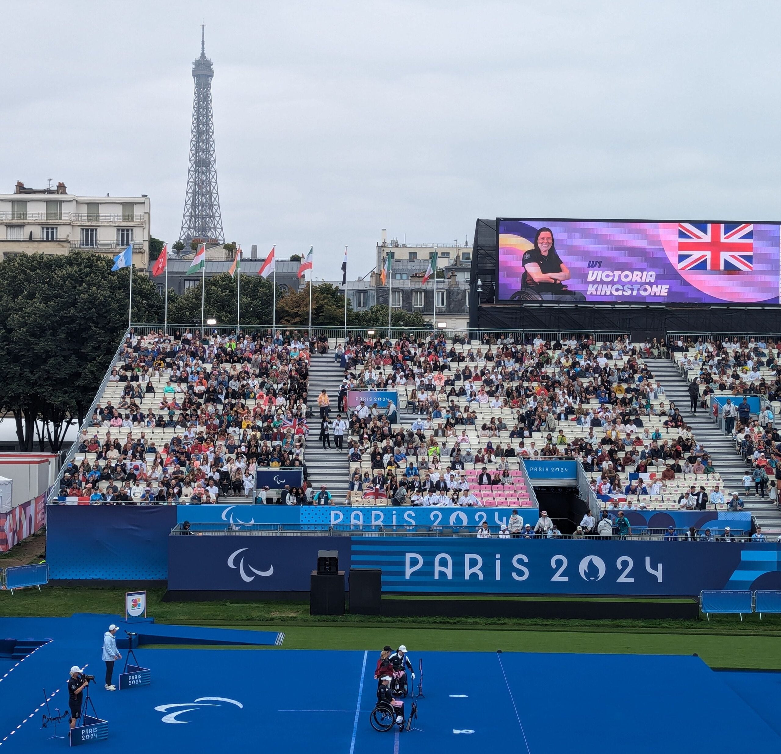 A wide shot of Paralympic archery event at Invalides in Paris 2024, with a large crowd seated in the stands. The Eiffel Tower is visible in the background, and a large screen displays the name and image of British archer Victoria Kingstone. The archers are positioned on a blue field, ready to compete.