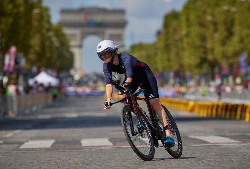 Claire Cashmore is pictured on her bike, leaning into a bend while wearing a GB kit and helmet. The Arc de Triomphe and cheering crowds line the road behind her creating a blurred backdrop.