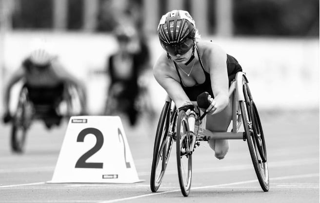 A black and white image of Olivia Gallagher, a wheelchair racer, competing on a track. She is in full racing form, with her arms propelling her racing wheelchair forward. The number 2 starting block is visible, indicating her lane. It captures a moment of intense athletic focus, representing the dedication and skill of para-athletes like Olivia