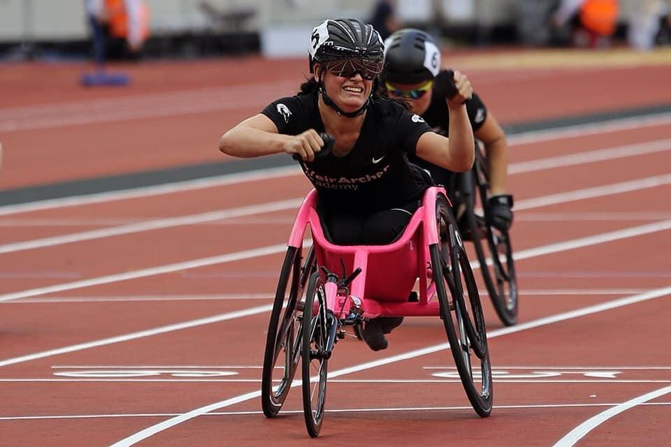 Olivia Gallagher is the main focus on a race track sat in her pink racing wheelchair wearing black sports kit and a helmet. She is smiling in a triumphant pose with both arms raised. There is another racer obscured from view behind her.