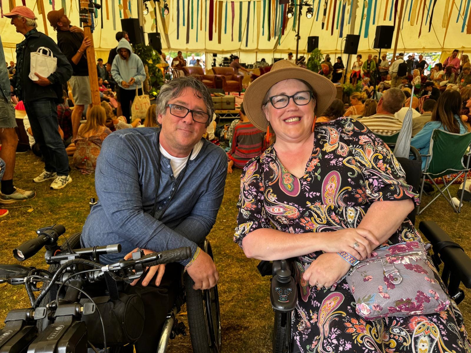 Grant Logan and Academy for Disabled Journalists alumna Yolanda Barker smiling at the camera inside a large festival tent. Both are seated in wheelchairs, surrounded by a lively audience at the Wilderness Festival. Colourful ribbons hang from the ceiling above a stage in the background.