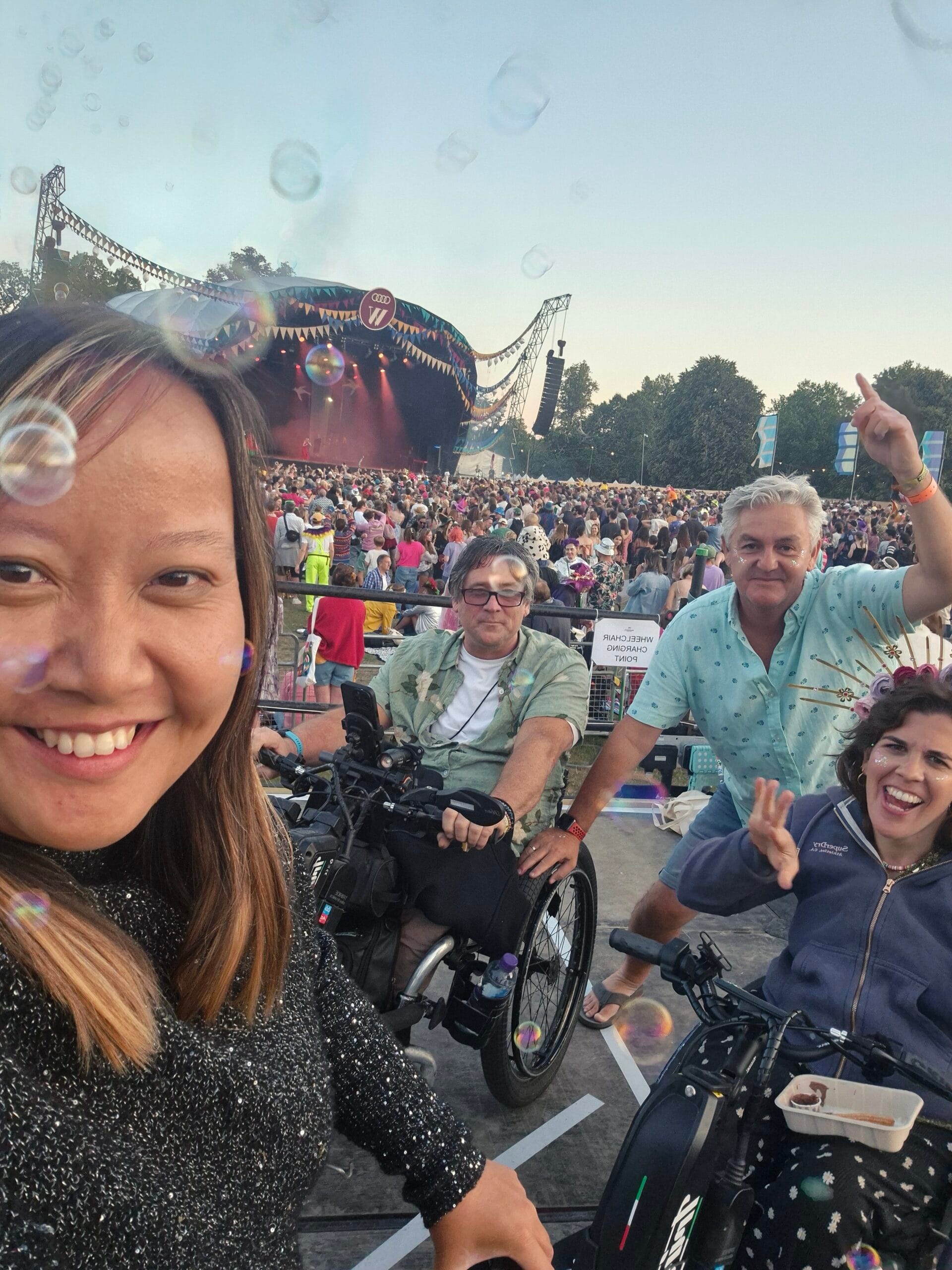 Group image of Kat, Sophie, Pete & Grant on the wheelchair platform behind a crowd of festival goers infront of the main stage with bubbles floating above them.