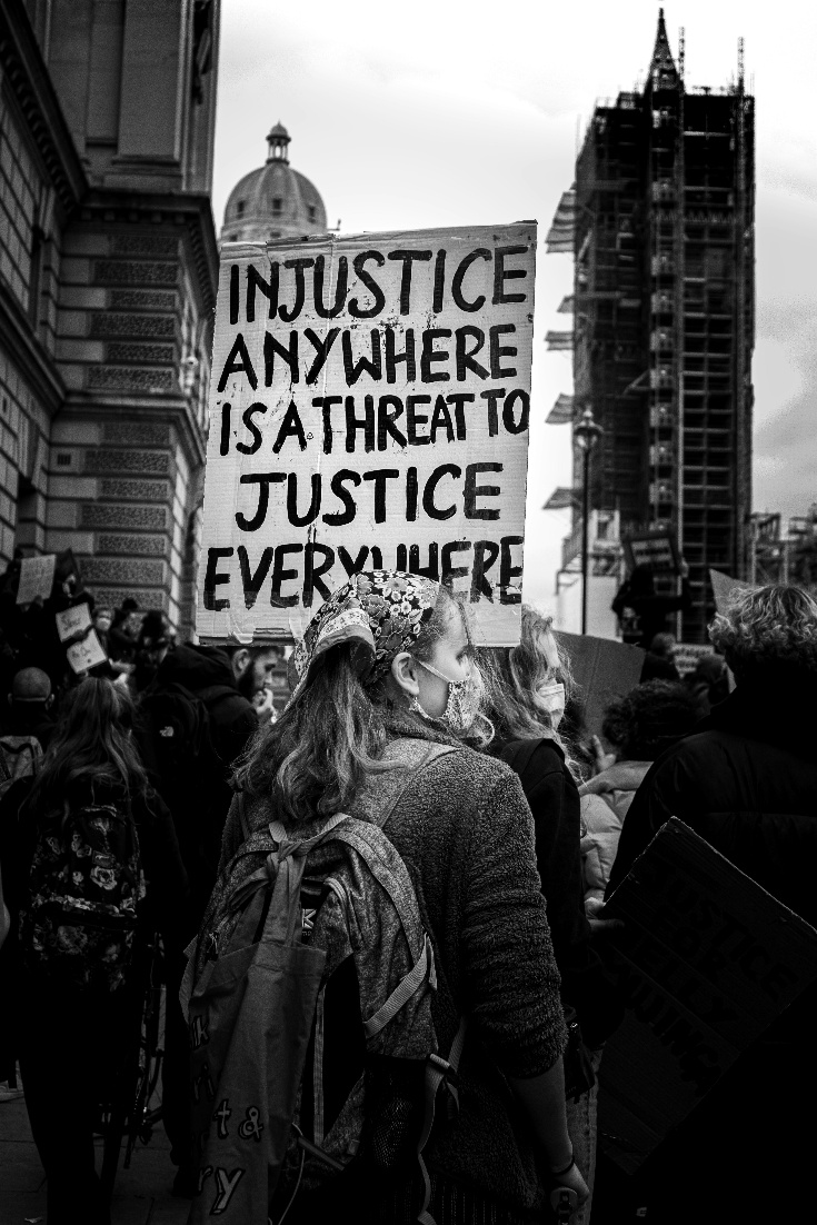 Black and white photo of  a street  protest with people wearing face masks holding placards.  the most prominent with the text 'Injustice anywhere is a threat to justice everywhere'