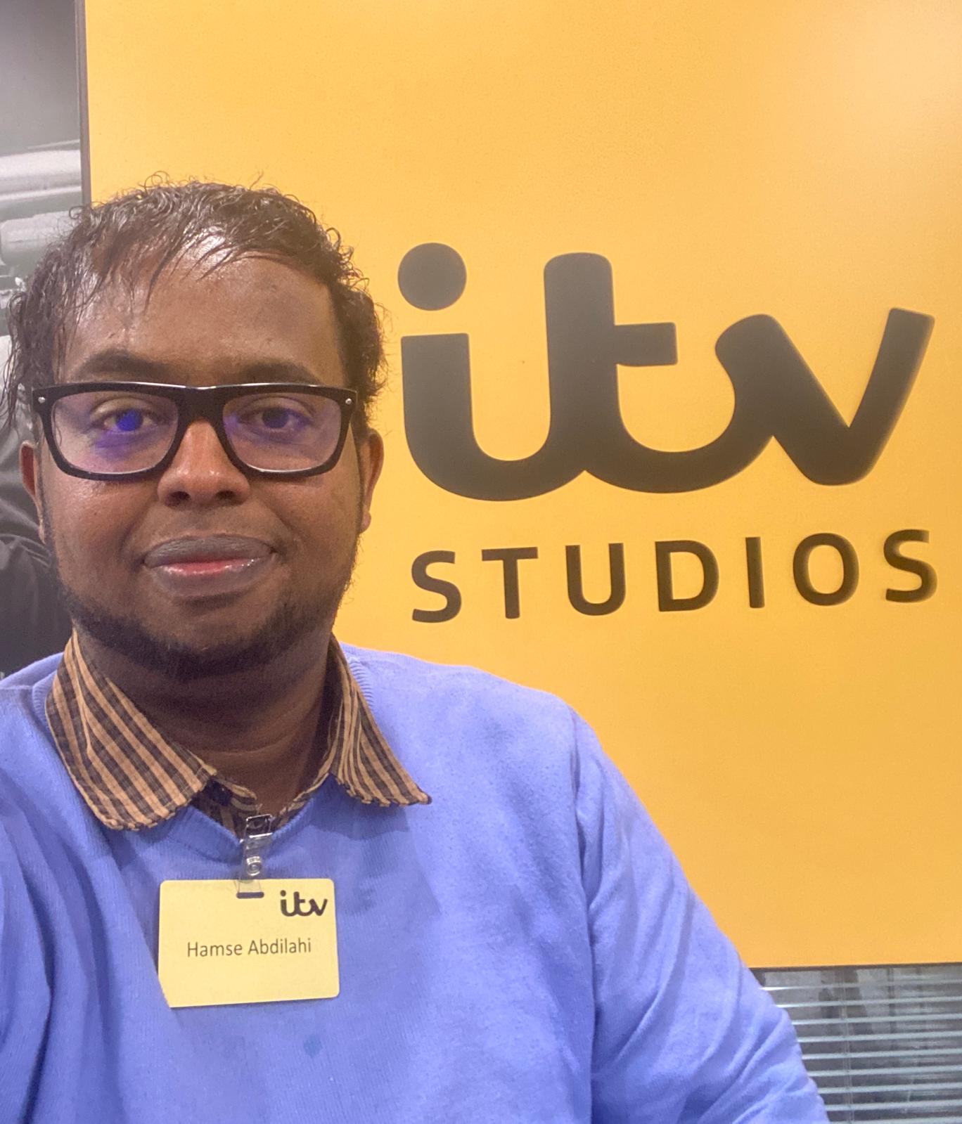 Image of Hamse smiling wearing glasses a purple jumper and shirt with an ITV studios sign in the background