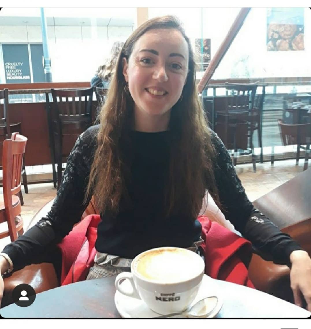 Nadia seated in a cafe smiling wearing a black long sleeved top with a cup of coffee on the table