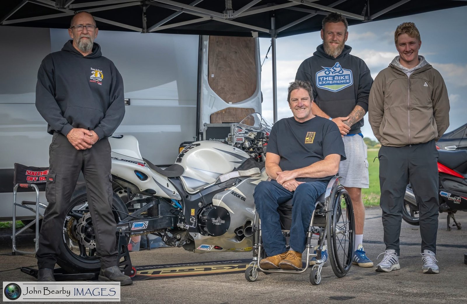 Talan Skeels-Piggins sits in his wheelchair beside a silver racing motorcycle, surrounded by three members of his racing and support team beneath a trackside canopy.