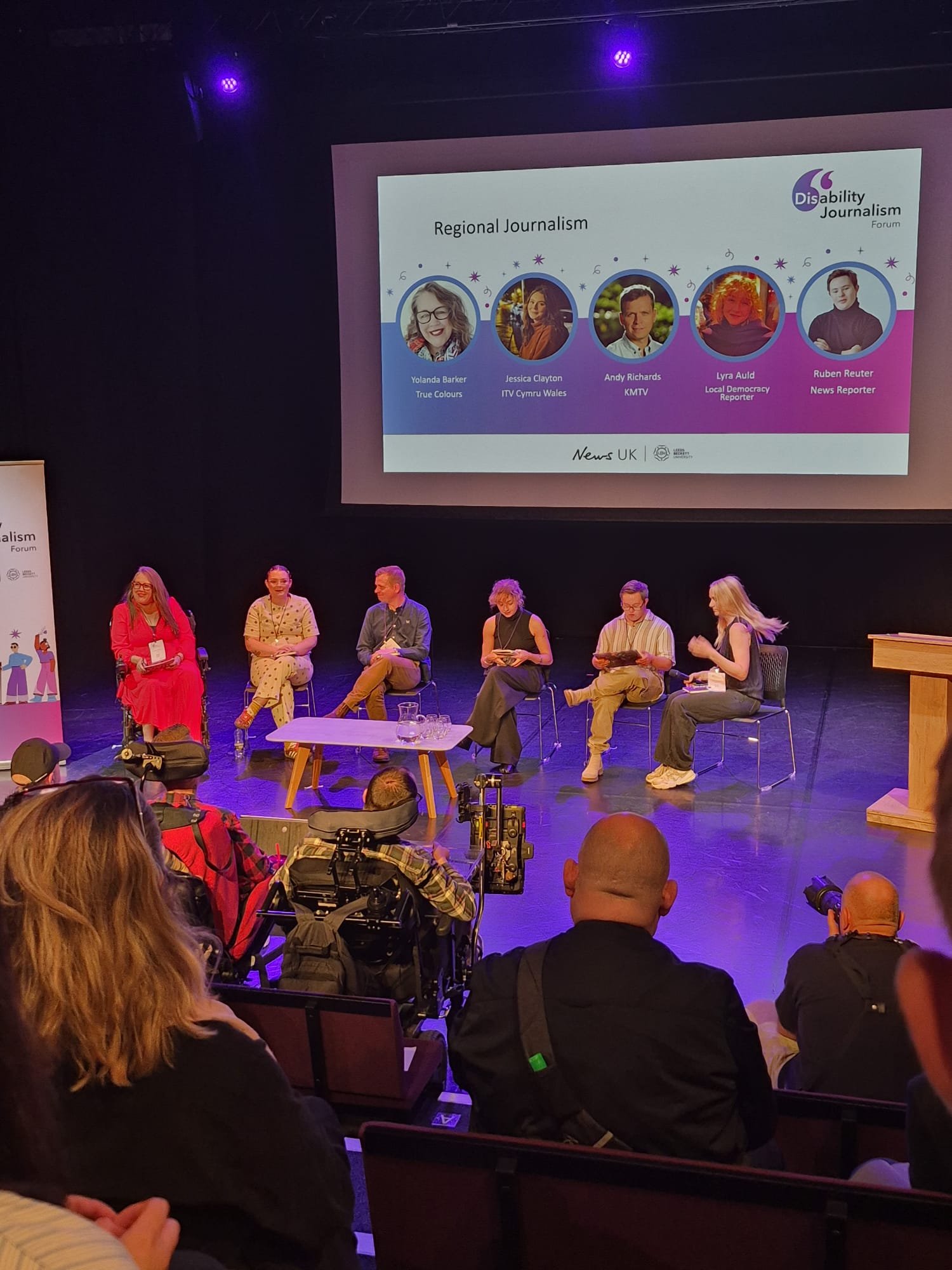 Yolanda Barker, seated on the left in red, moderates the Regional Journalism panel at the Disability Journalism Forum Conference. Fellow panellists are seated beside her on stage, with their names and photographs displayed on a large screen above.